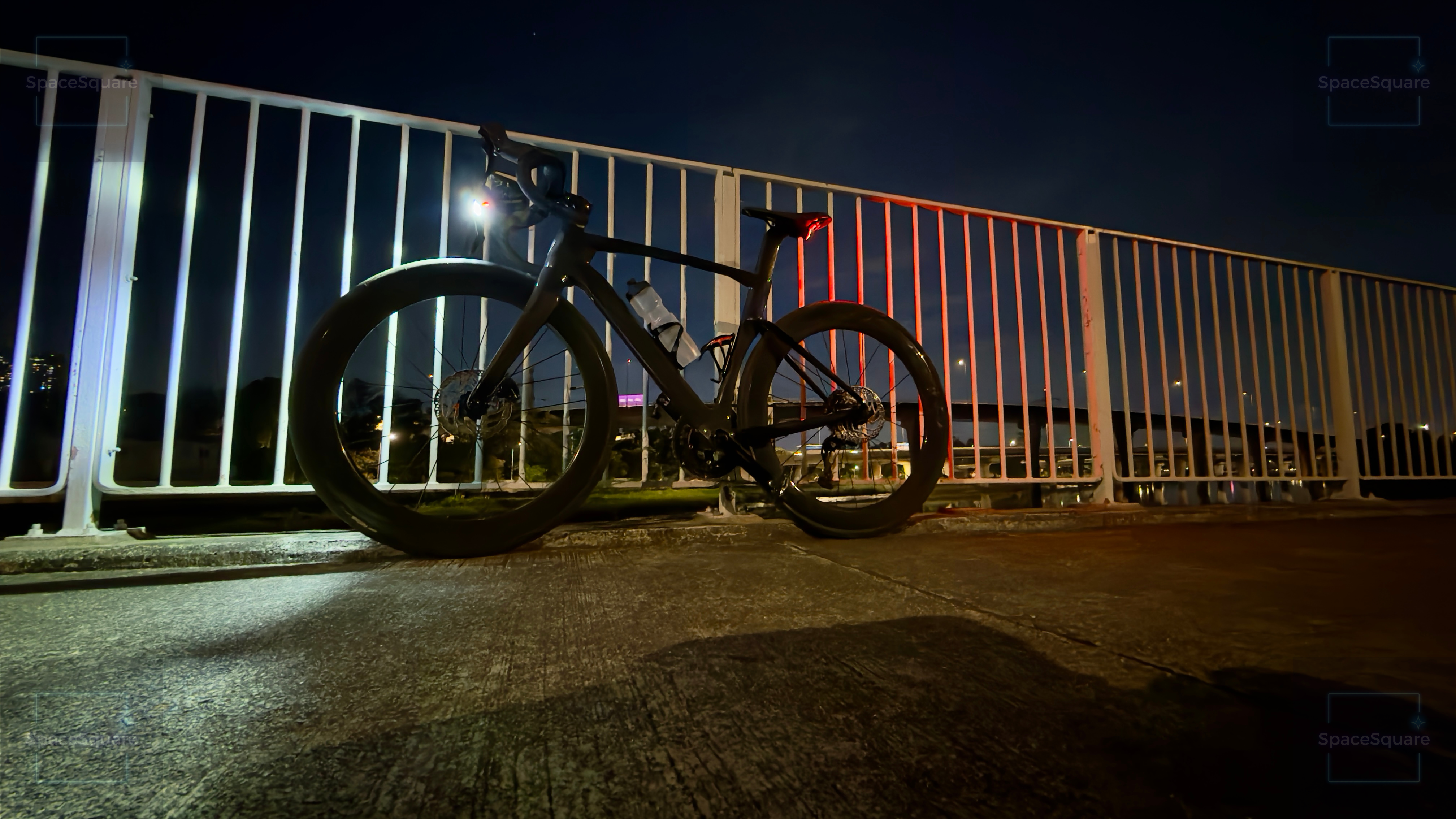 Road bike leaning against a white railing on a bridge over Kam Tin River, Nam Sang Wai, Yuen Long — front light glowing white, rear light glowing red, Hong Kong skyline in the distance